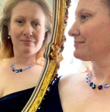 Woman looking in mirror wearing a silver necklace with nine 20 mm octagon shaped crystals that ombre in color from a deep ocean blue to a celestial lavender. The earrings match the center necklace crystal that is the deep extra sparkly blue.